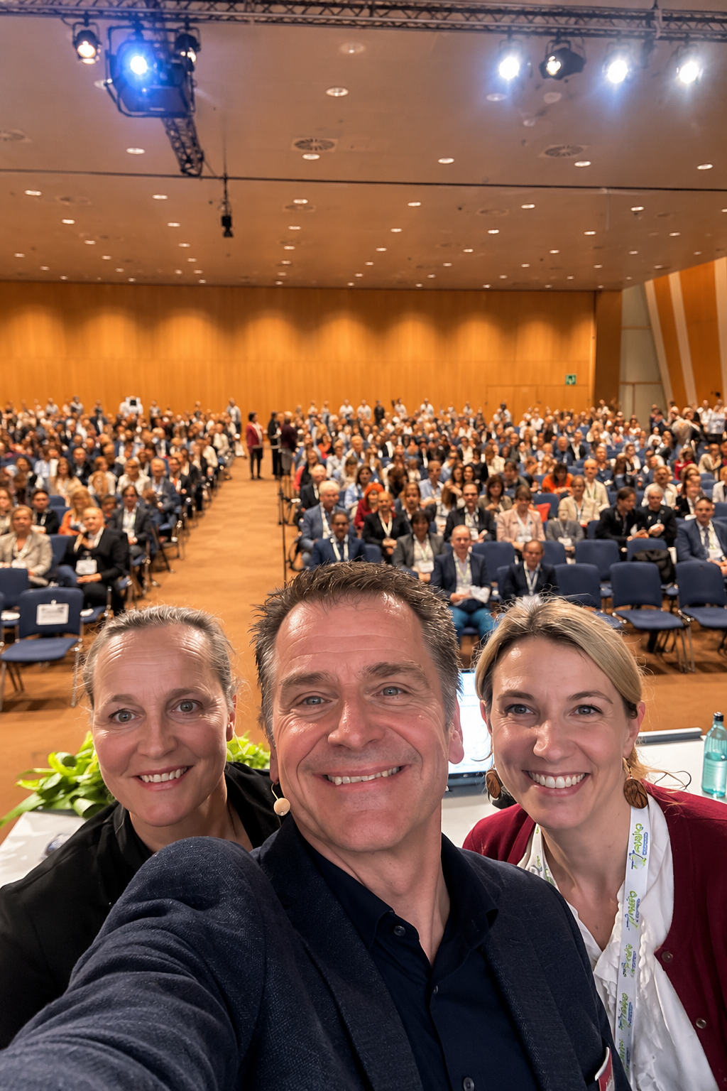 Patrick van Oirschot with two fellow healthcare professionals at an international hospital pharmacy conference, with a large audience in the background.